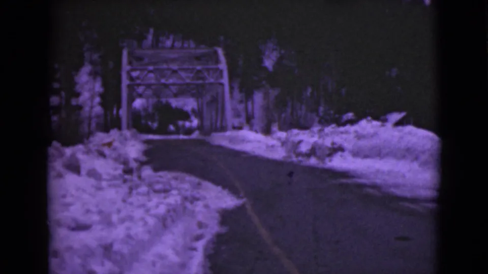 The forward view down a snowy Alaska road shows high plowed banks, dark pavement, roadside trees, and a metal bridge ahead.
