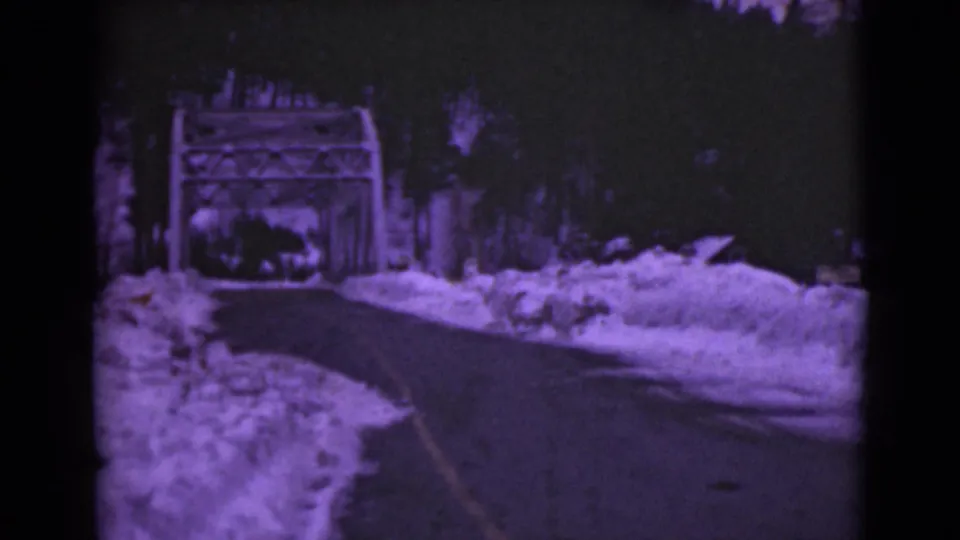 A soft 8mm frame shows the road curving through heavy snow toward the same bridge, with trees and film-edge darkening visible.