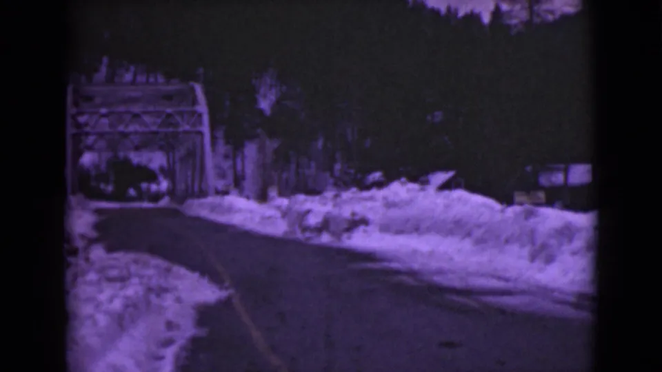 The final frame keeps the metal bridge ahead as the road and snowbanks fill the foreground, with a purple archival film cast.