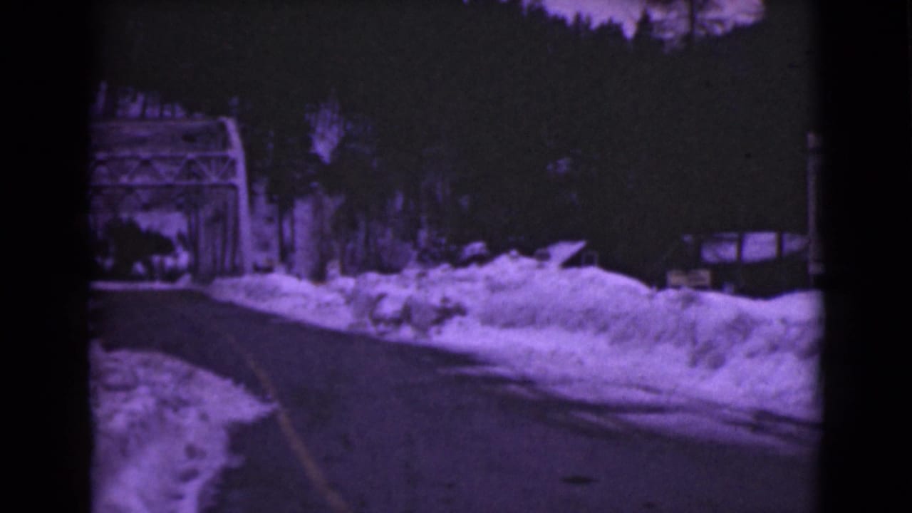 1946 Alaska 8mm frame showing a dark road bordered by deep snow, with a bridge structure and trees in the background.