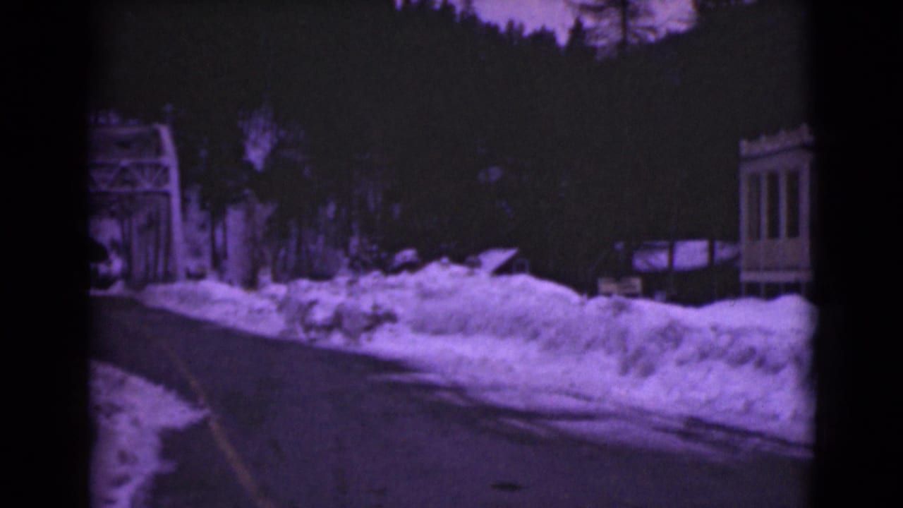 Snowbank-lined road with bridge structure at left and a small building at right.