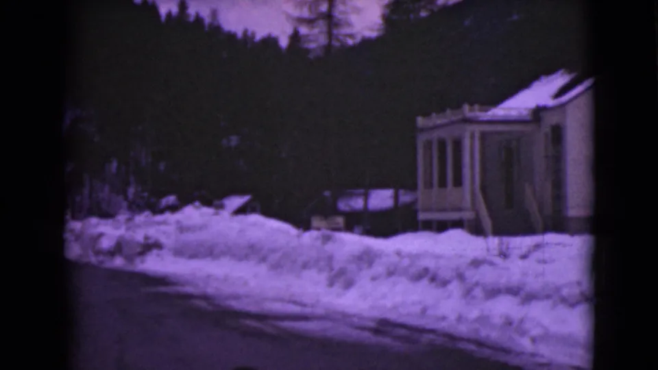 1946 Alaska home-movie frame with a wooden roadside house, deep snowbanks, and a dark tree-covered backdrop.
