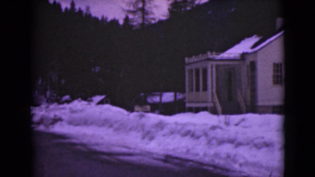 1946 Alaska home-movie frame of a white house near high snowbanks, with dark trees and a hillside in the background.