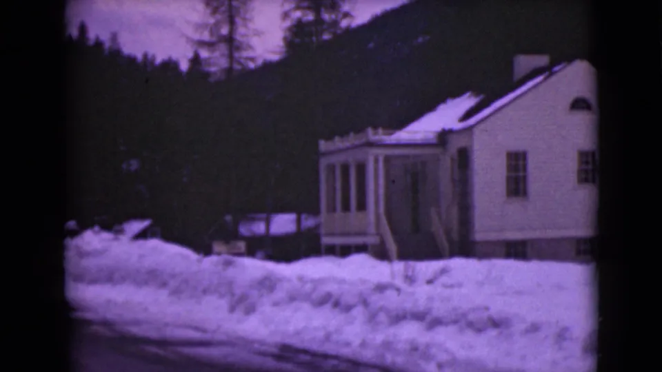 1946 Alaska frame showing a house exterior, porch, roadside snowbanks, trees, and a dark hillside under winter light.