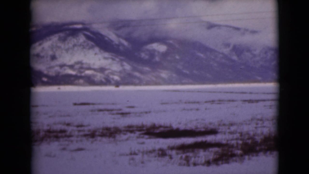 1946 Alaska 8mm frame showing snow-covered mountains across a frozen tundra or lake, with dark exposed patches in the foreground.