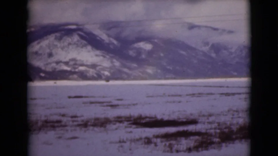 1946 Alaska home-movie frame with a snowy mountain ridge, pale frozen ground or water, and dark tundra patches across the lower frame.