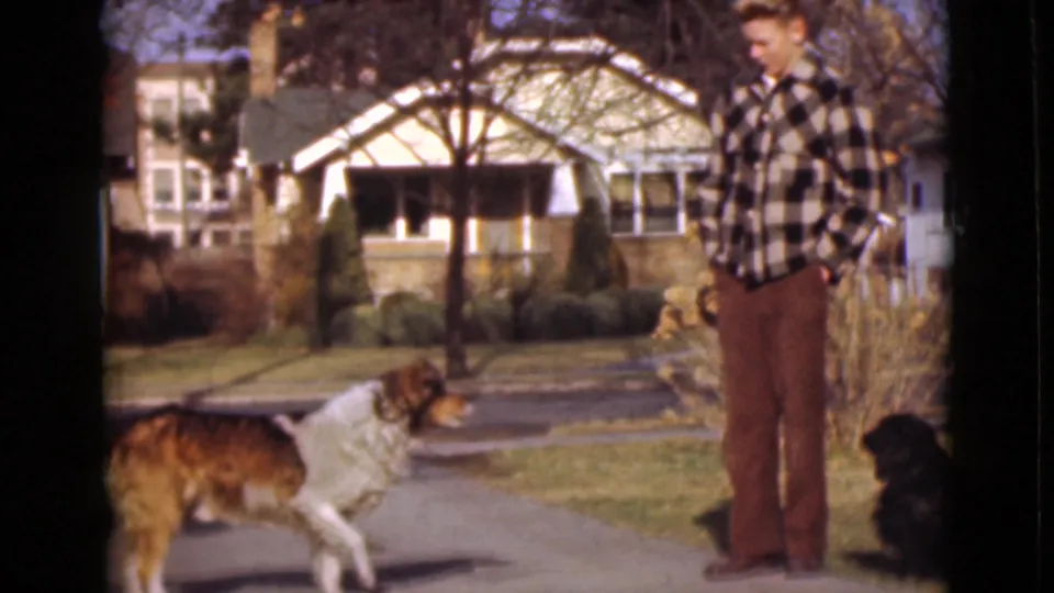 1946 Alaska home-movie frame with Fred on the sidewalk, the larger dog turned toward him, and the smaller black dog sitting nearby.