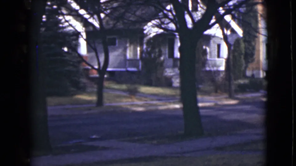 A 1946 Alaska home-movie view of a still residential block, with an empty road, sidewalks, lawns, and houses behind leafless trees.