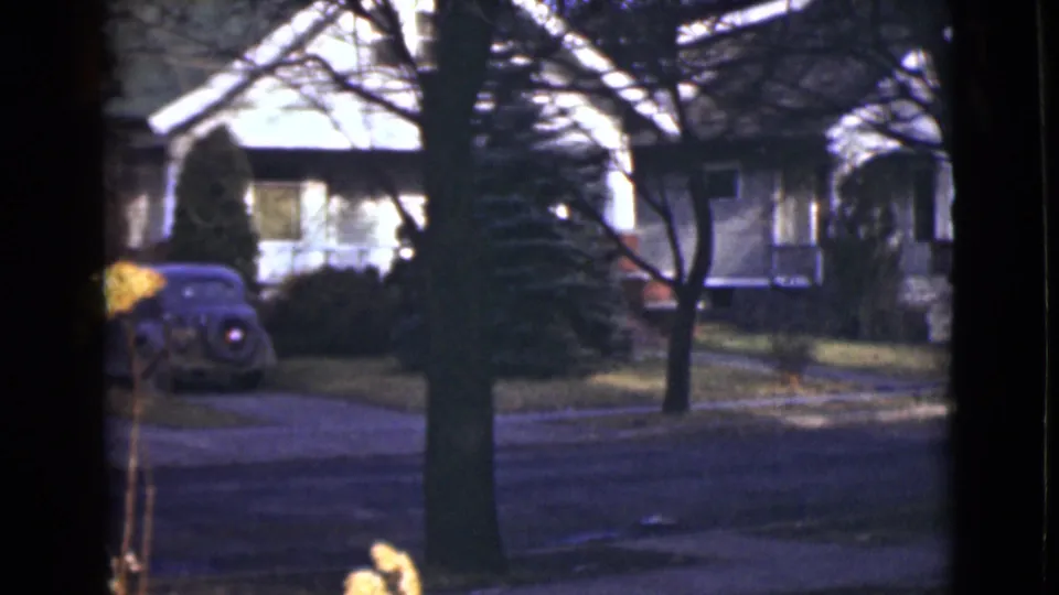1946 Alaska home-movie frame with a rounded car partly visible near a driveway, framed by trees and modest houses.