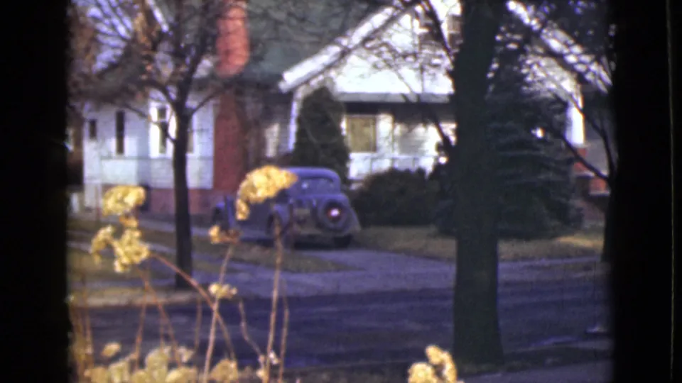 1946 Alaska home-movie frame with a parked rounded car near small houses, seen through trees from across the street.