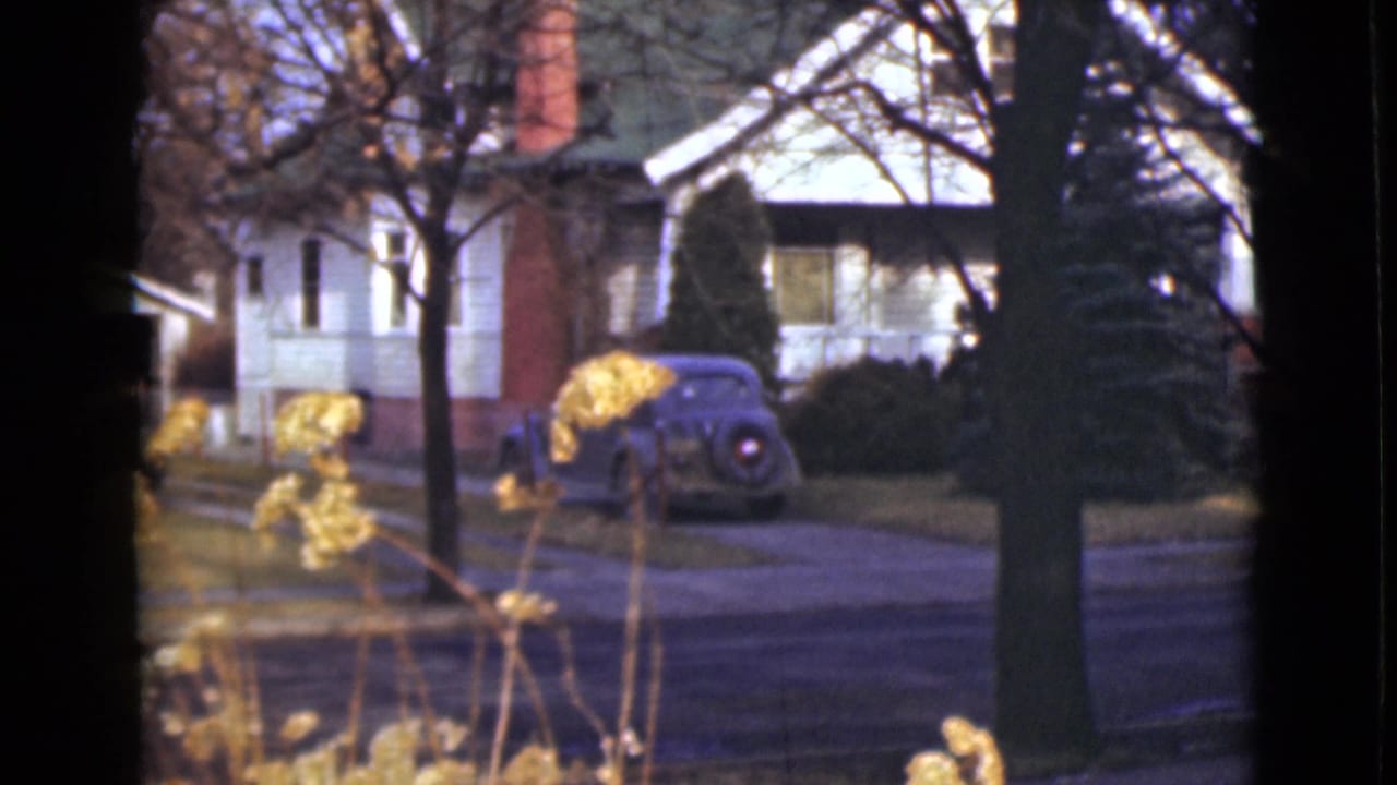 1946 Alaska 8mm frame showing a white residential house, brick chimney, blue parked sedan, bare trees, and yellow plants in the foreground.