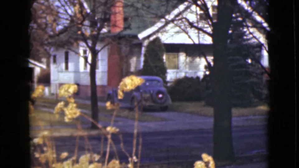 1946 Alaska 8mm frame showing a white residential house, brick chimney, blue parked sedan, bare trees, and yellow plants in the foreground.