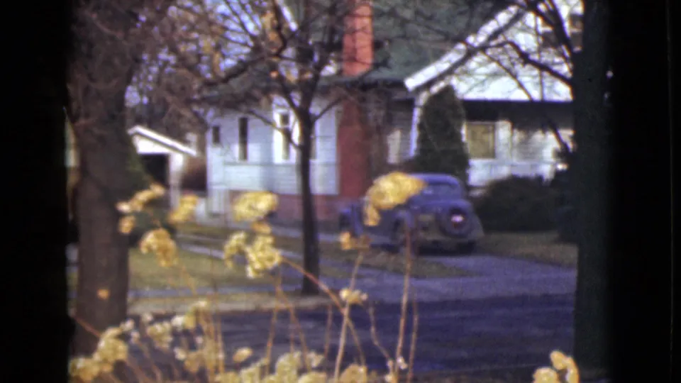 1946 Alaska 8mm view of a modest house with a brick chimney and blue sedan, partially obscured by trees and plants.