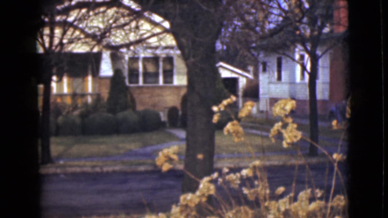 1946 Alaska 8mm frame showing a brick residential house across a quiet road, partly screened by trees and foreground flowers.