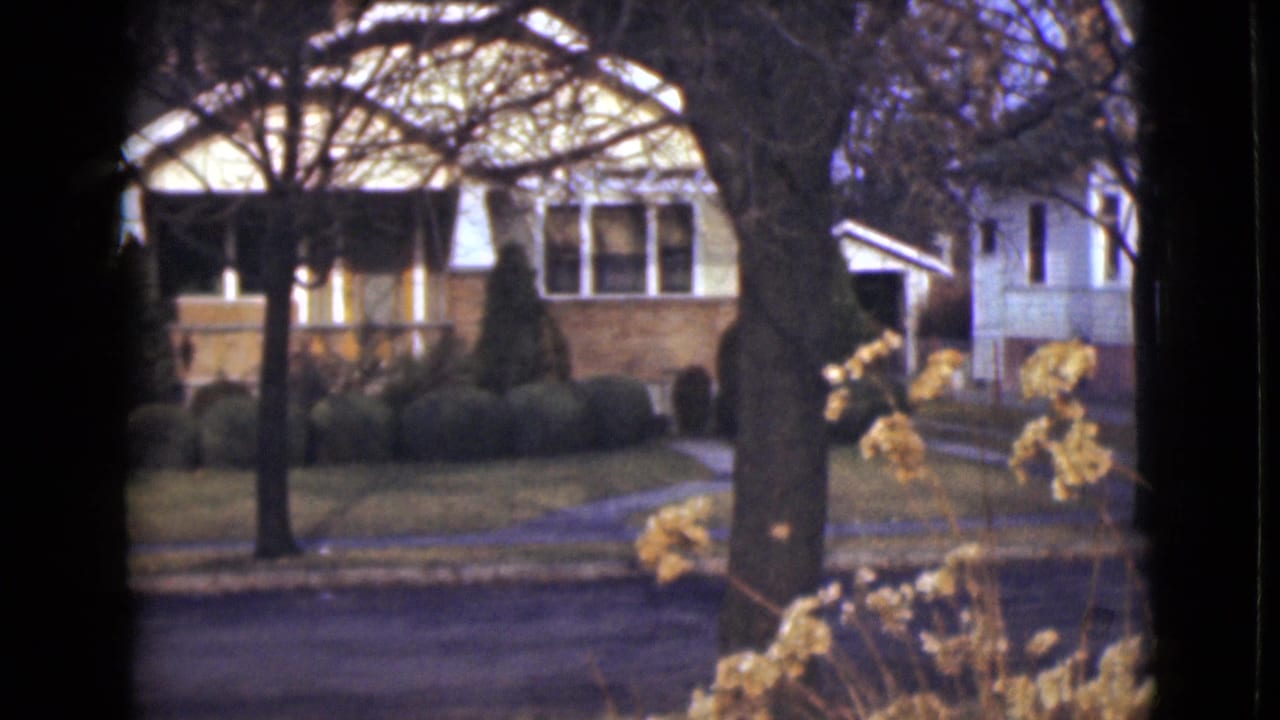 1946 Alaska home-movie frame of a quiet residential frontage with a porch, trimmed shrubs, trees, and road.