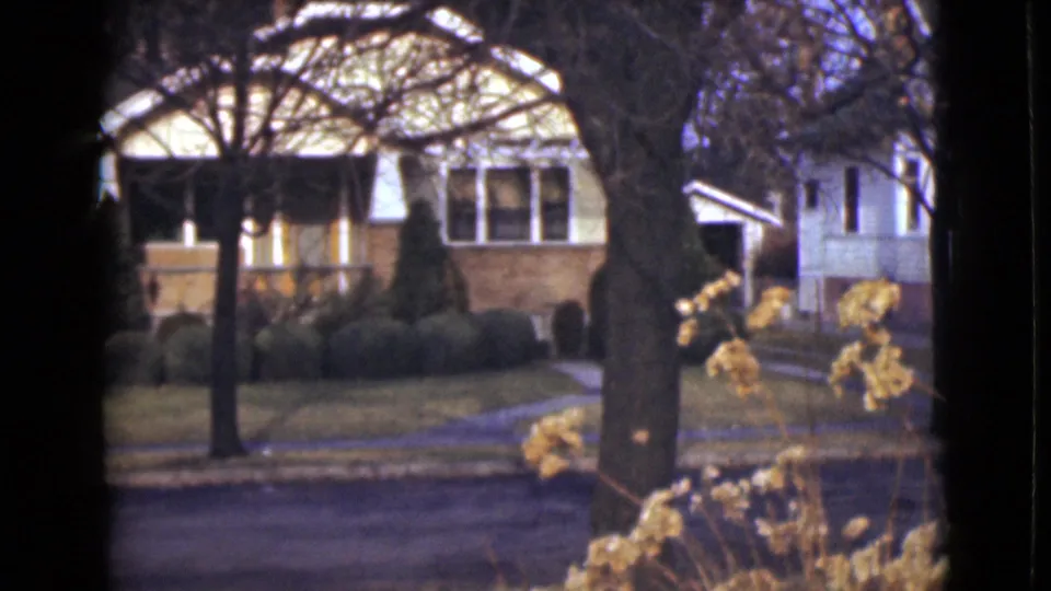1946 Alaska home-movie frame of a quiet residential frontage with a porch, trimmed shrubs, trees, and road.