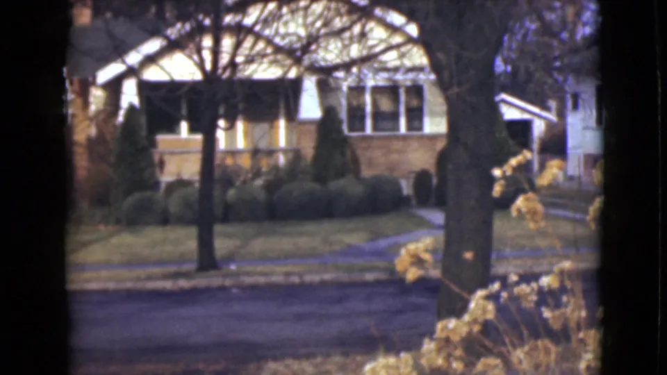 1946 Alaska 8mm frame showing a residential yard and house across the road, with winter-bare trees and dry flowers nearby.