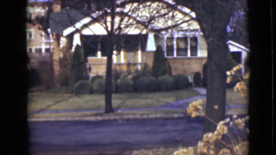1946 Alaska home-movie frame of a house and landscaped yard across a quiet street, framed by trees and dry flowers.