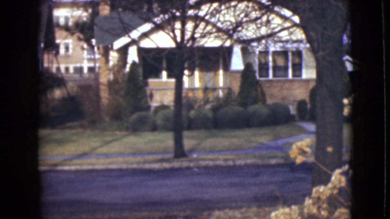 1946 Alaska home-movie frame showing a detached house across an empty street, with leafless trees, clipped shrubs, lawn, and sidewalk.