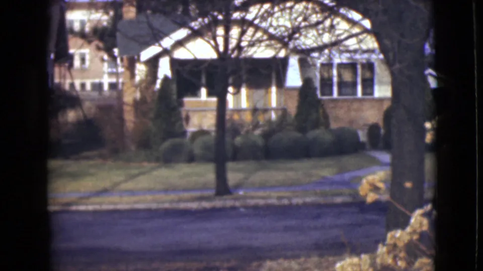 1946 Alaska home-movie frame showing a detached house across an empty street, with leafless trees, clipped shrubs, lawn, and sidewalk.