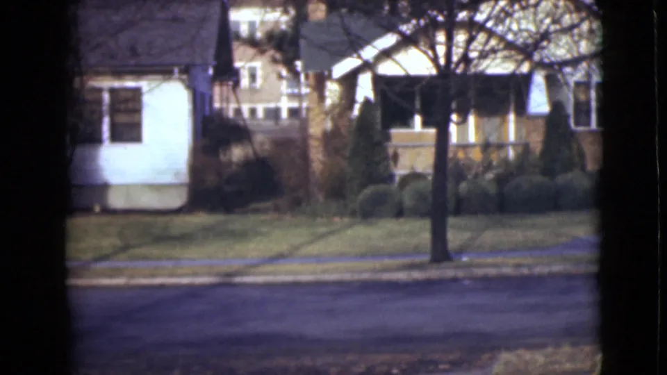 1946 Alaska home-movie frame with neighboring house fronts, a dark tree trunk, bare branches, lawn, shrubs, sidewalk, and no visible people.