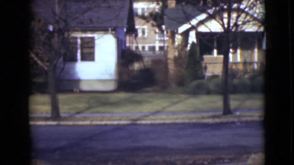1946 Alaska suburban street frame showing house exteriors, leafless trees, clipped shrubs, a sidewalk, and an empty paved road.
