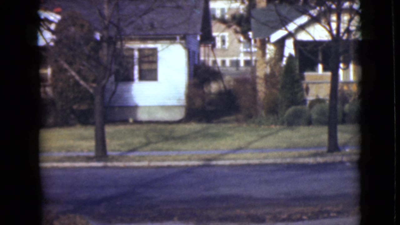 1946 Alaska home-movie frame showing small houses, lawns, bare trees, and a paved residential road.