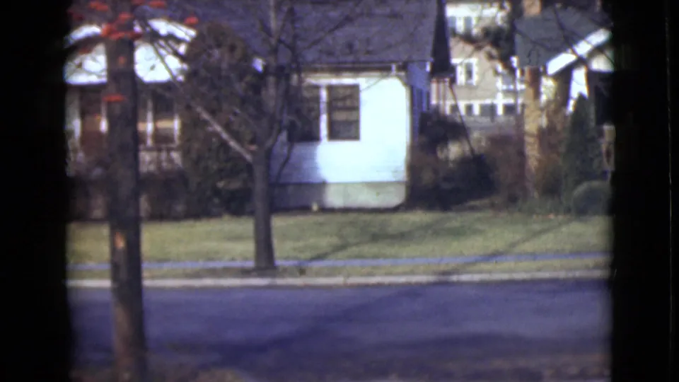 1946 Alaska residential view with modest houses, bare trees, lawns, and a paved road in soft 8mm color.