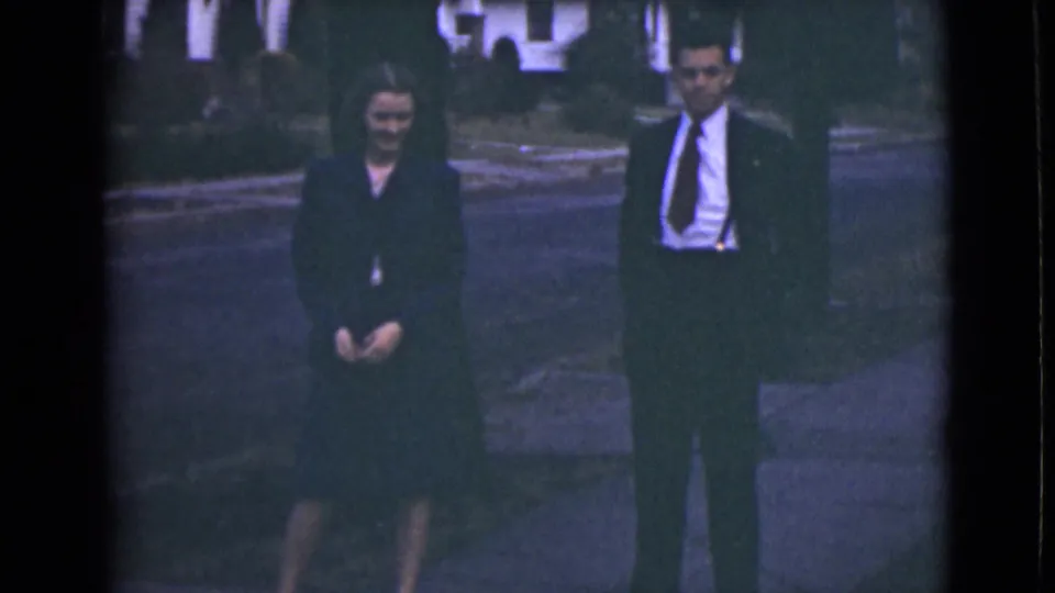 1946 Alaska home-movie frame showing a woman in dark clothing and a suited man standing on a neighborhood sidewalk.
