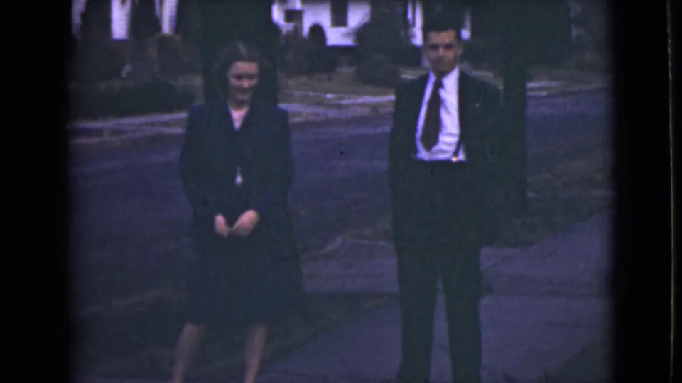 1946 Alaska 8mm frame with two adults standing on a sidewalk, houses and street receding behind them.