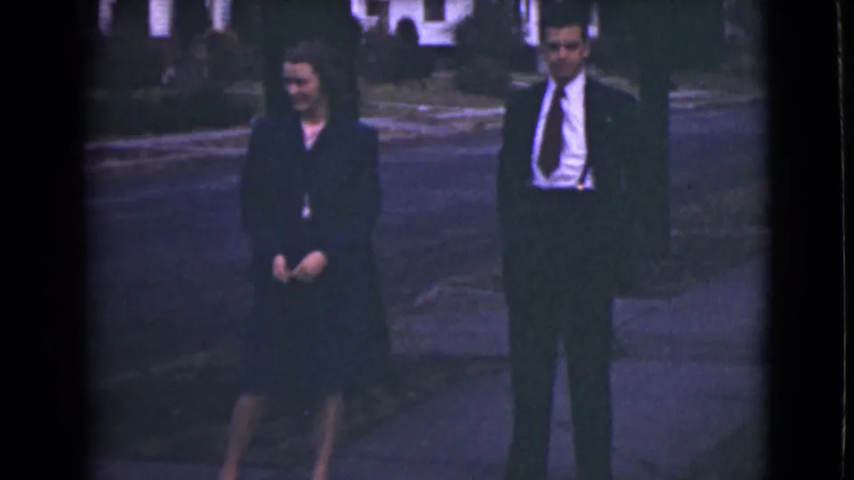 In this 1946 Alaska home-movie frame, a well-dressed woman and man stand apart on a sidewalk in soft daylight.