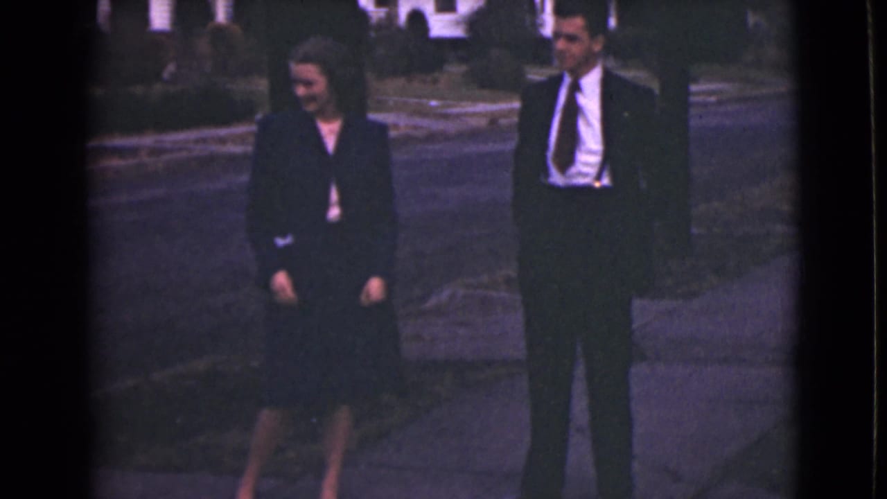 In 1946 Alaska home-movie footage, a woman and man stand on a sidewalk near a curb in dark formal clothing.