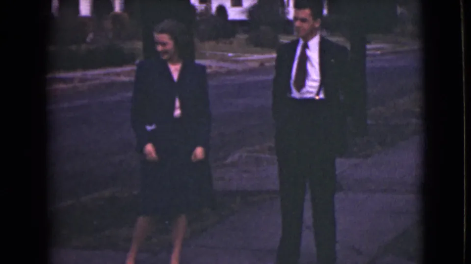 In 1946 Alaska home-movie footage, a woman and man stand on a sidewalk near a curb in dark formal clothing.