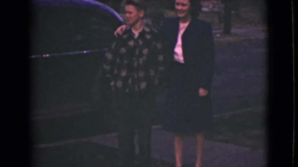 Near a parked car, the patterned-shirt man places an arm around the suited man while the woman stands close by.