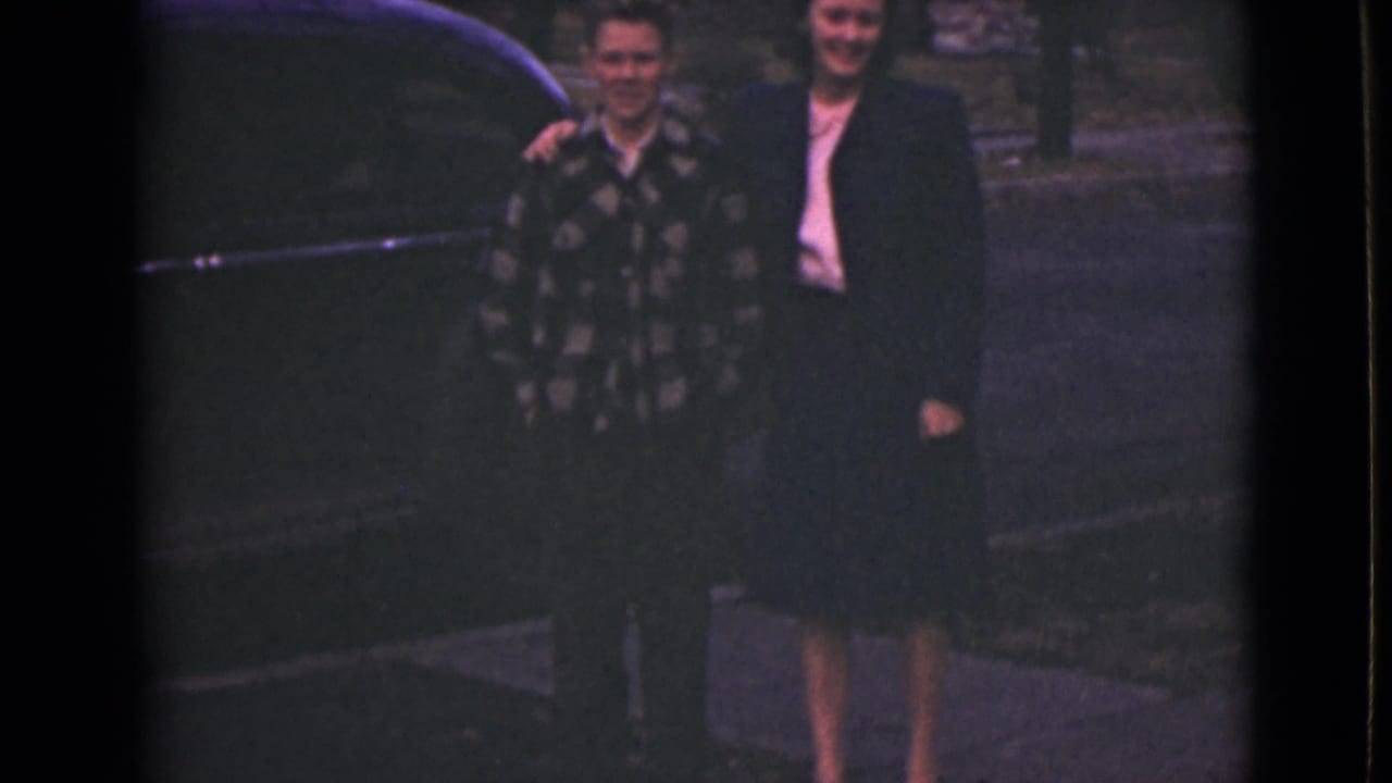 Two people smiling beside a dark vintage car on a paved outdoor surface.