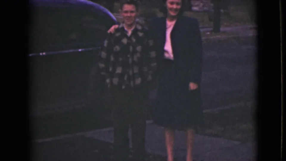 In 1946 Alaska home-movie footage, a son in a patterned shirt stands beside a woman in a dark suit near a dark car.