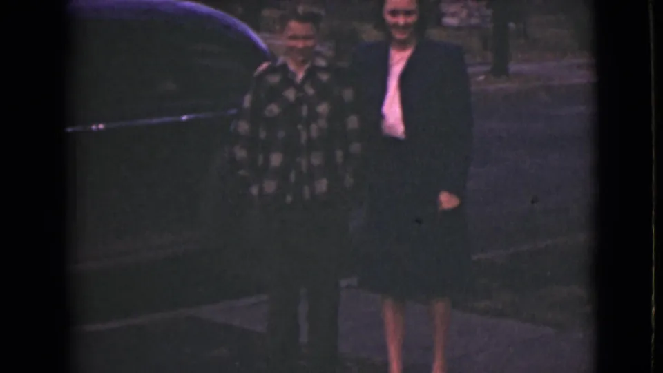 A woman in a dark suit and a younger male in a patterned shirt smile near a car in metadata-supported 1946 Alaska footage.