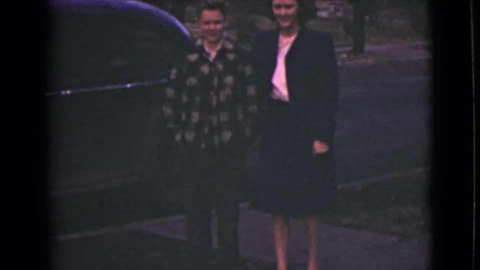 The 8mm frame shows a mother and son smiling beside a dark car, with faded color and outdoor pavement visible.