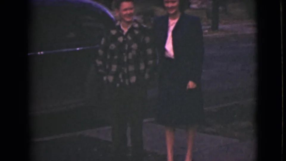 A 1946 Alaska home-movie frame shows a mother and son standing together beside a dark vintage car outdoors.