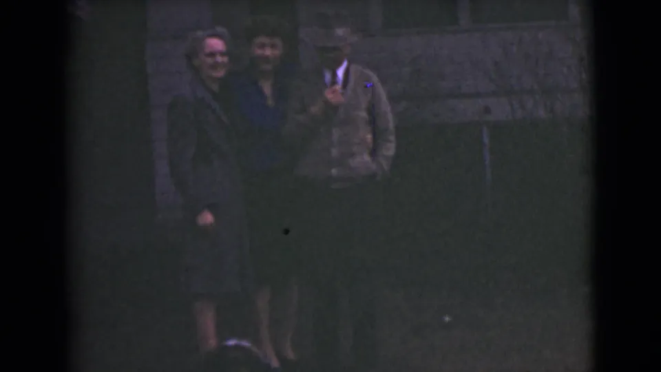 1946 Alaska home-movie frame with two women and a man in a hat smiling together outside a house.