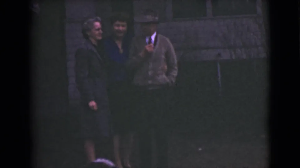 1946 Alaska home-movie frame showing two women and a man grouped on a lawn beside a house exterior.