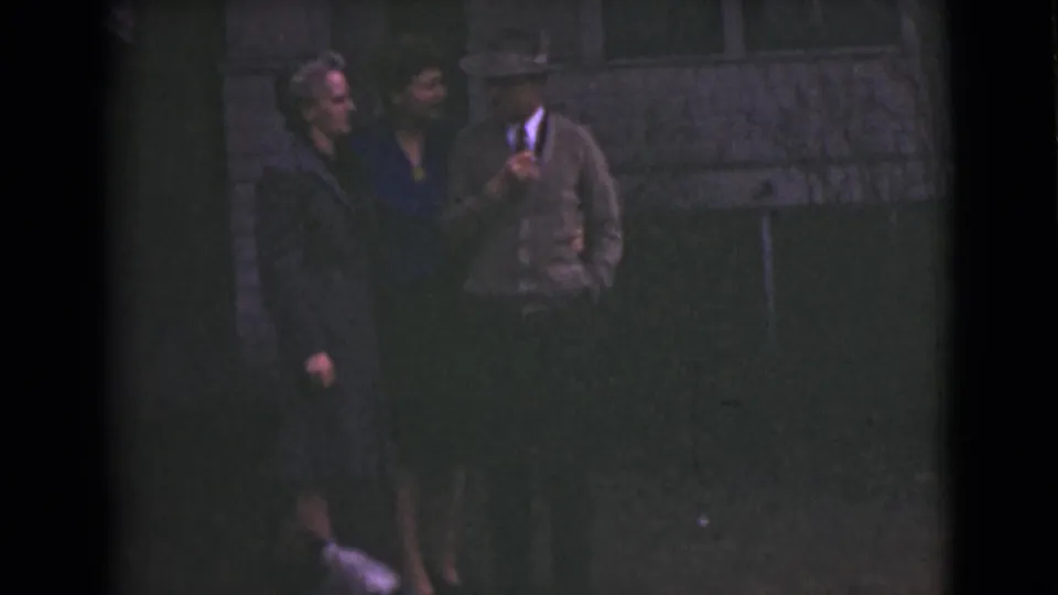 1946 Alaska home-movie frame with three adults outside a house and a dog partly visible near their feet.