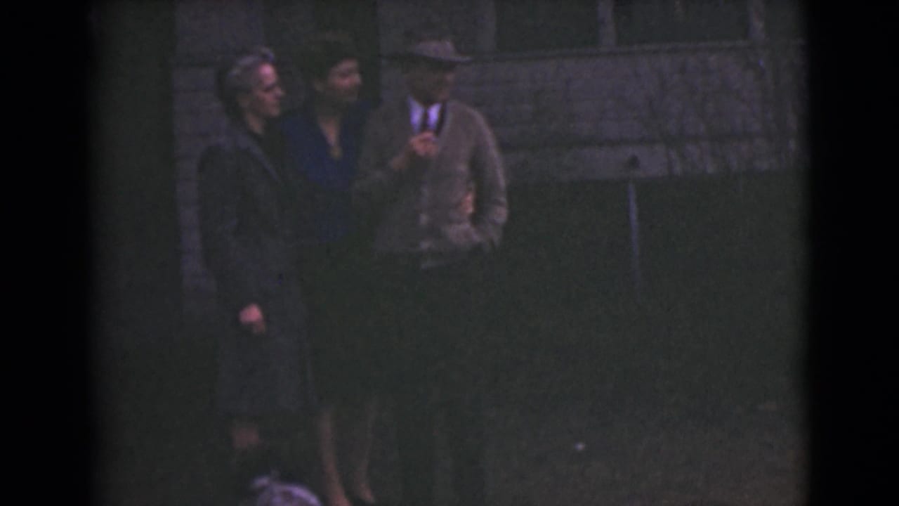 Three adults stand on grass outside a stone building, with one person holding a hat.