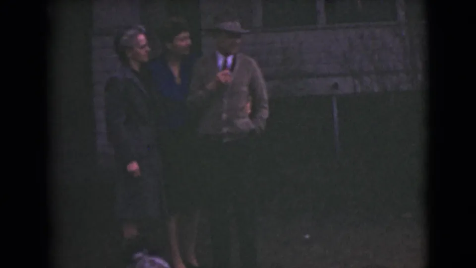 Three adults in 1946 Alaska home-movie footage stand outside on grass near a stone wall, appearing to pose for the camera.
