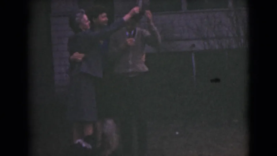 In a dim 1946 Alaska 8mm frame, three adults stand together while one woman lifts her arm in a playful gesture.