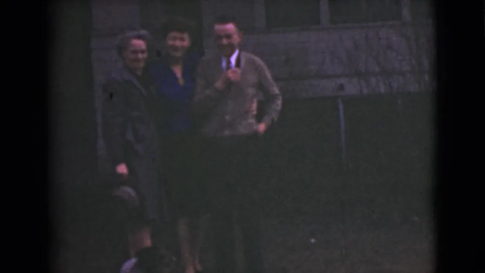 Three adults smile toward the camera in 1946 Alaska home-movie footage, with a small dog partly visible near their feet.