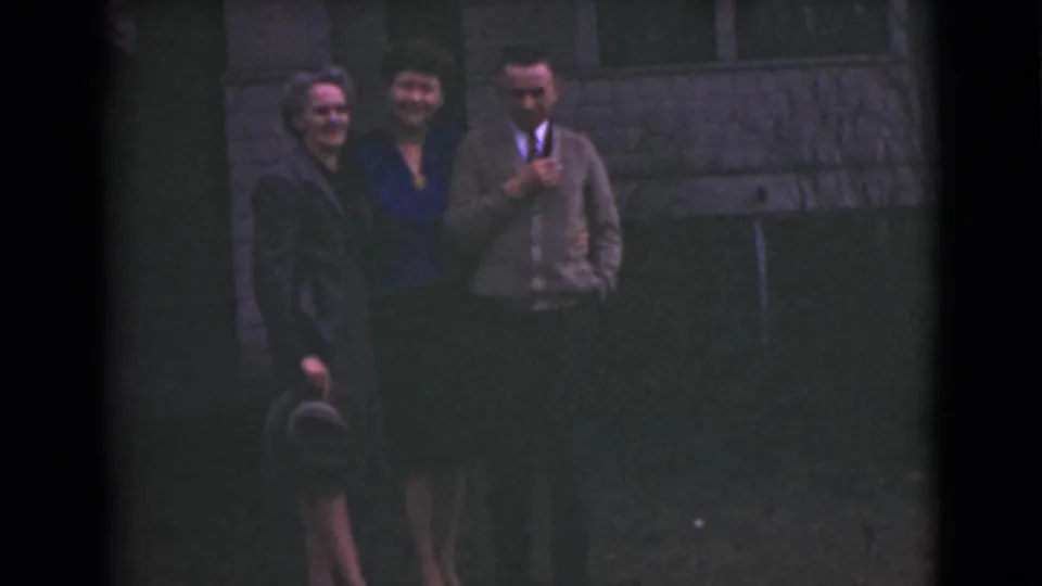 Three adults in Alaska, 1946, stand close together and smile for the 8mm camera outside a stone building.