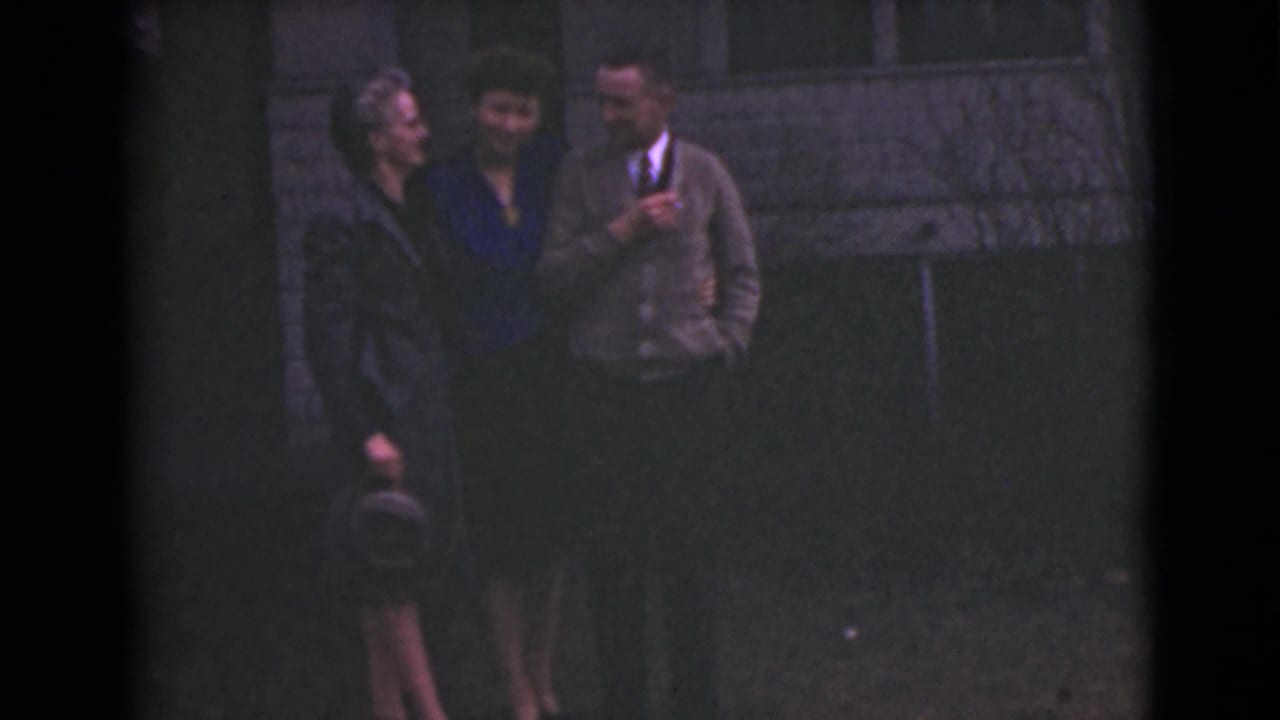 Three young adults stand outside by a wall, with one woman holding a small hat.