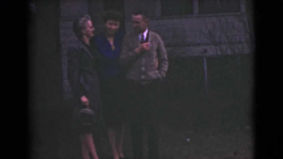 In 1946 Alaska home-movie footage, two women and a man in a cardigan and tie stand close together outside against a wall.