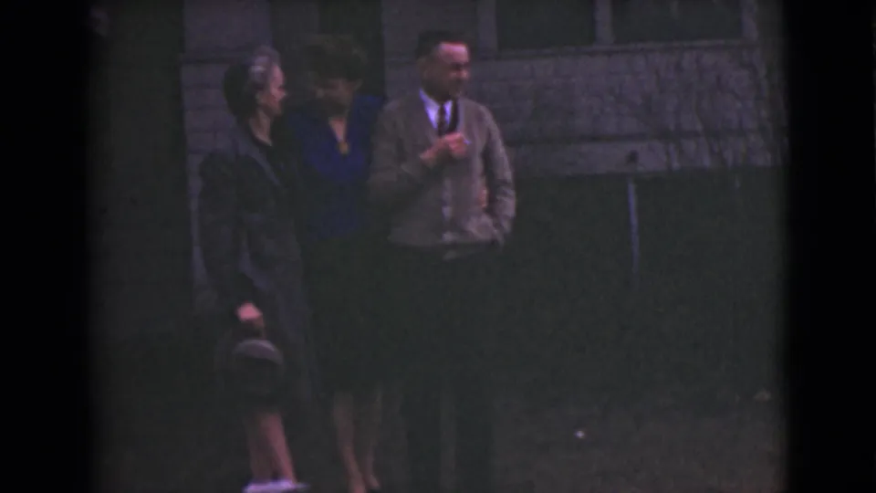 The group remains near the wall in grainy 8mm footage, with the man looking toward the women as they stand arm in arm.